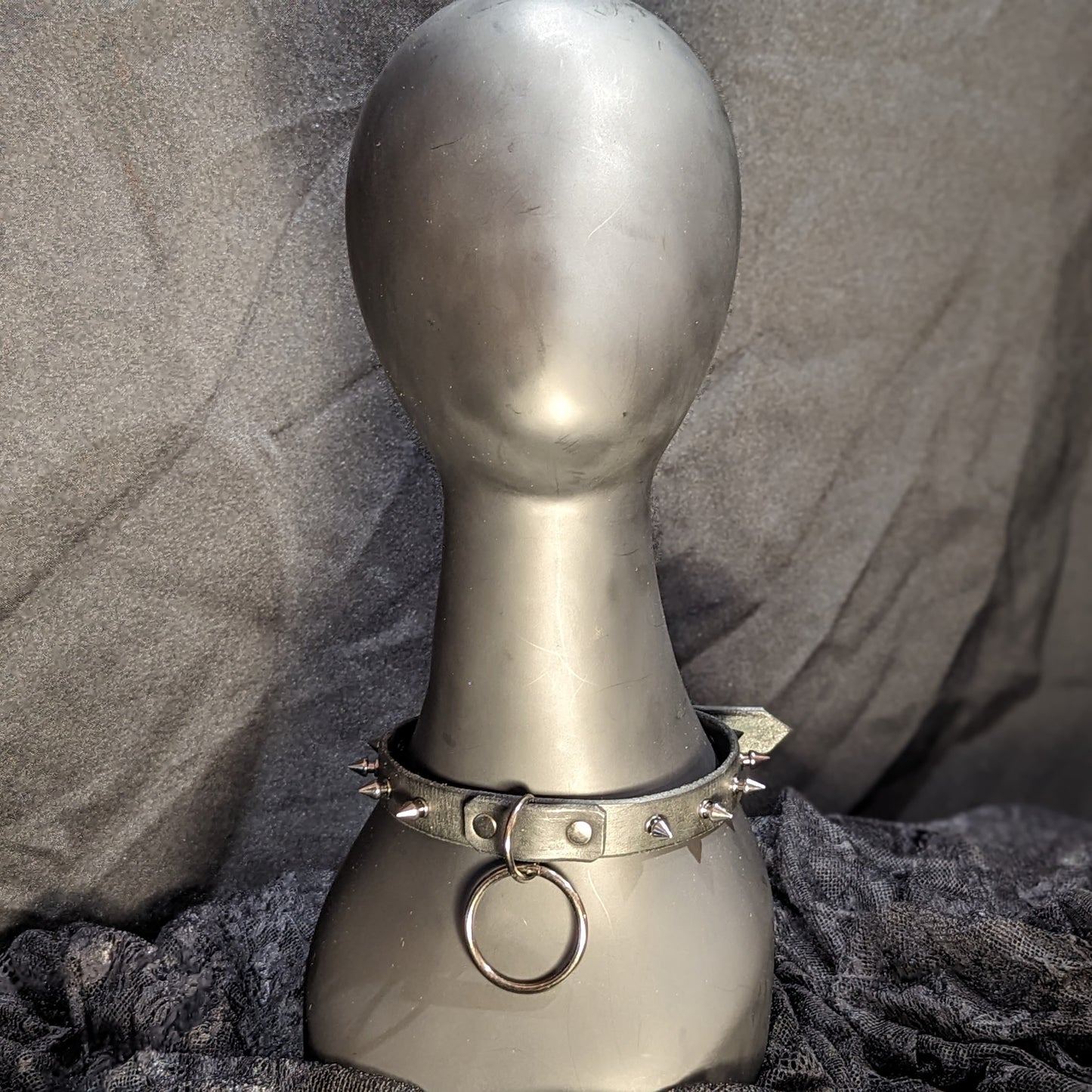 Black leather collar by Smell My Leather. This collar has small 1/4" gunmetal spikes and a dangling 1.5" O-ring. mid range image. The collar is displayed on a black plastic mannequin.