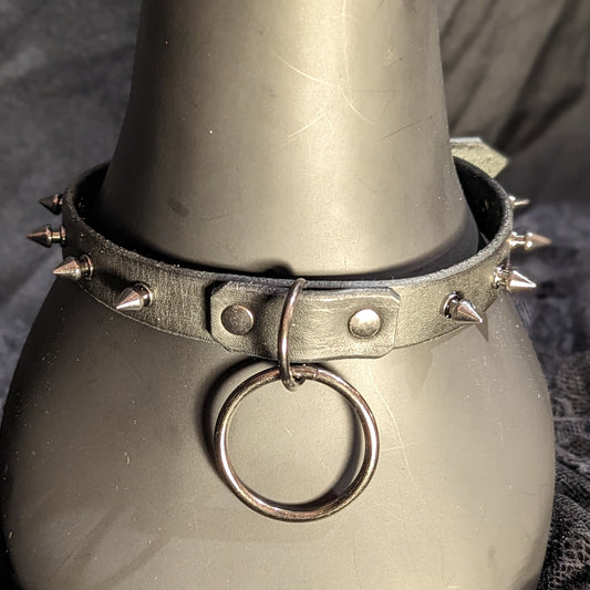 Black leather collar by Smell My Leather. This collar has small 1/4" gunmetal spikes and a dangling 1.5" O-ring. Close up image. The collar is displayed on a black plastic mannequin.